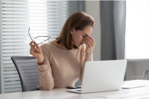 Women pinching bridge of her nose, glasses in one hand, looking down at laptop on her table.