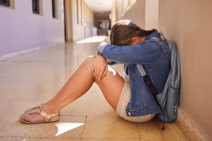 Teenage girl on the ground in a school hallway with her head on her knees face hidden.