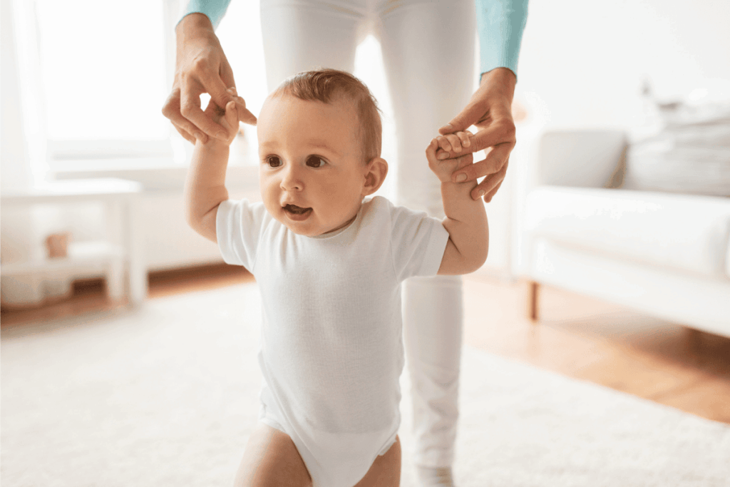 Picture of a toddler learning to walk, arms held by mother in the background.