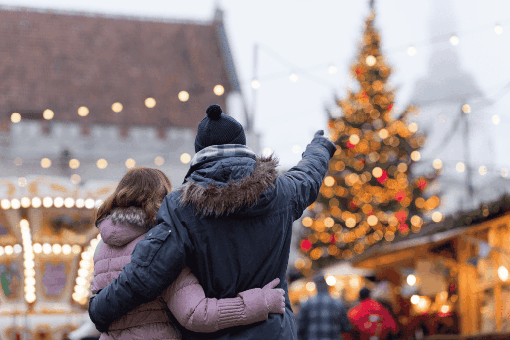A parent and teen standing together at an outdoor Christmas market, looking at holiday lights and a decorated tree.
