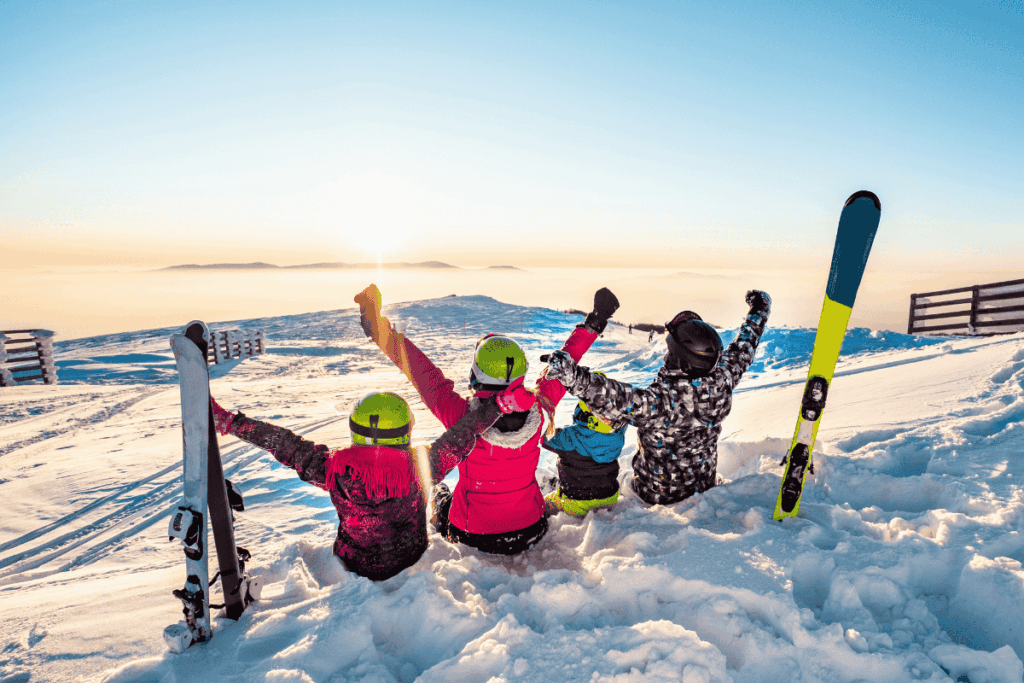 A family sitting together on a snowy mountain at sunrise, with skis beside them, sharing a joyful winter moment.