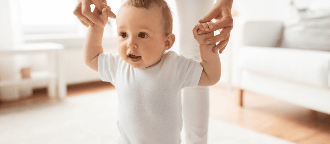 Picture of a toddler learning to walk, arms held by mother in the background.