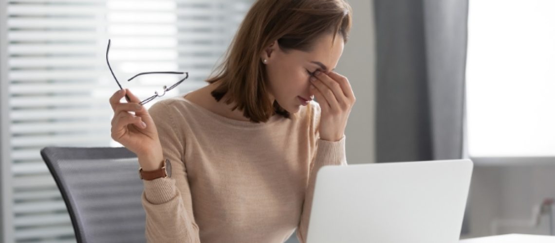 Women pinching bridge of her nose, glasses in one hand, looking down at laptop on her table.